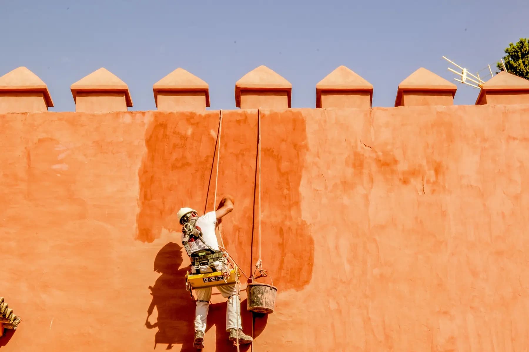 Un artisan suspendu à des cordes enduit un haut mur ocre à créneaux, sous un ciel bleu